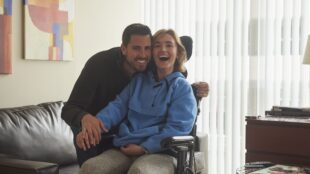 a woman in a wheelchair is smiling. she is wearing a blue hoodie and gray sweatpants. a man to her right is seen embracing her and smiling with her.