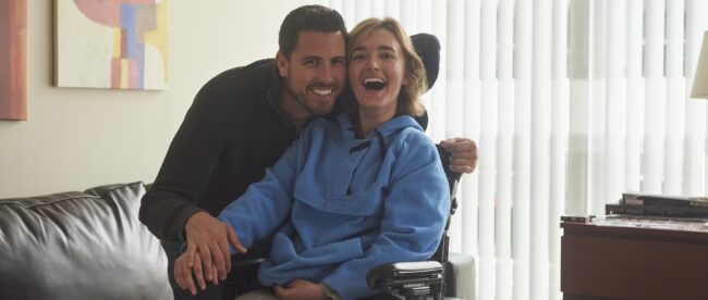 a woman in a wheelchair is smiling. she is wearing a blue hoodie and gray sweatpants. a man to her right is seen embracing her and smiling with her.