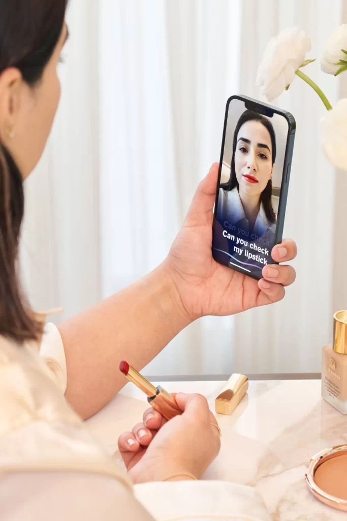 A woman is holding a lipstick in her right hand and a phone in her left. The phone has the VMA app open with the text "can you check my lipstick" on it, implying that the user is asking VMA app for feedback about the lipstick she jus applied.