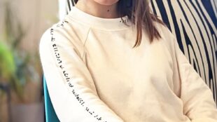 A woman wearing a cream colored sweater. Braille on sleeve reads: this sweater was made alongside the blind and visually impaired community. Braille on neckline reads: aille design