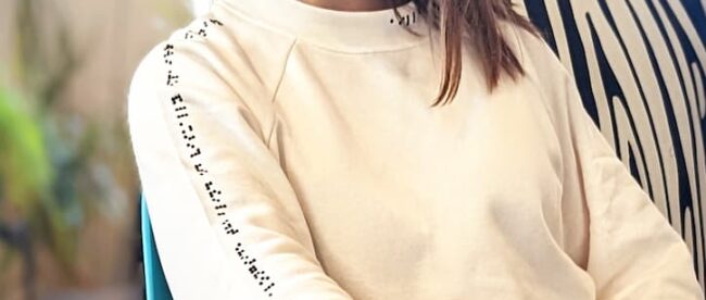 A woman wearing a cream colored sweater. Braille on sleeve reads: this sweater was made alongside the blind and visually impaired community. Braille on neckline reads: aille design