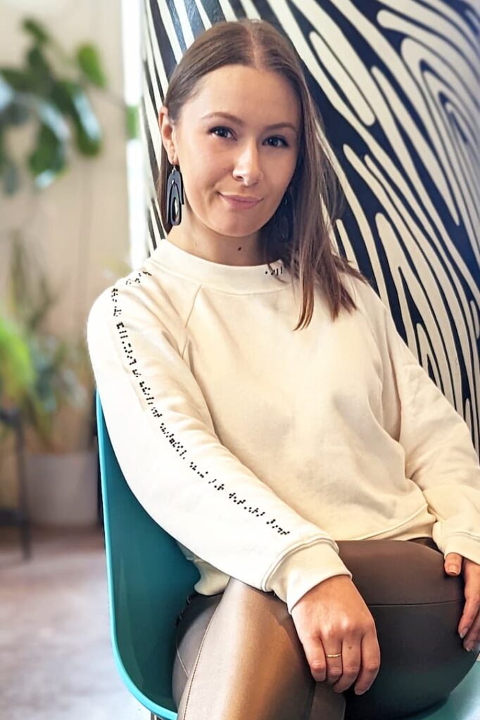 A woman wearing a cream colored sweater. Braille on sleeve reads: this sweater was made alongside the blind and visually impaired community. Braille on neckline reads: aille design