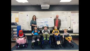 5 children in wheelchair for whom the funds were raised for an accessible playground pose for a photo in a classroom. Behind them are a white male and female holding an enlarged check for $10,000 made to glen lake elementary.