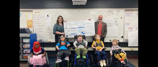 5 children in wheelchair for whom the funds were raised for an accessible playground pose for a photo in a classroom. Behind them are a white male and female holding an enlarged check for $10,000 made to glen lake elementary.