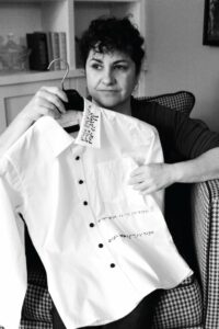 A black and white photo of a woman holding the first prototype created by Alexa. It’s a white shirt with braille beadwork on the left color and middle of the shirt.