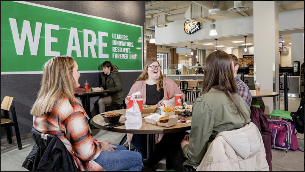 Four students sitting in a cafeteria and chatting while eating fast food. Behind them is a banner that says “we are leaders, innovators, resilient, forever UND”.