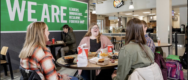 Four students sitting in a cafeteria and chatting while eating fast food. Behind them is a banner that says “we are leaders, innovators, resilient, forever UND”.