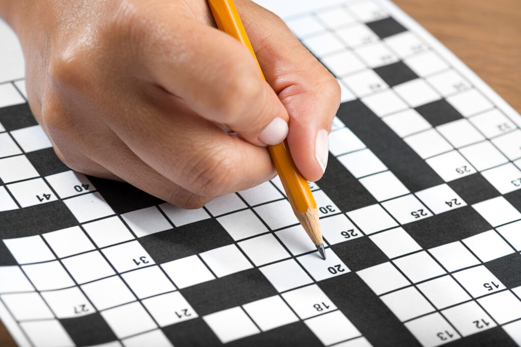 extreme close up of someone's hand. They are holding a pencil and filling up clues on a crossword puzzle.