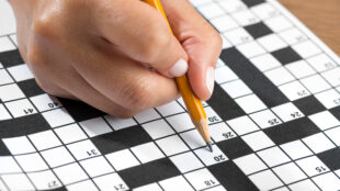 extreme close up of someone's hand. They are holding a pencil and filling up clues on a crossword puzzle.