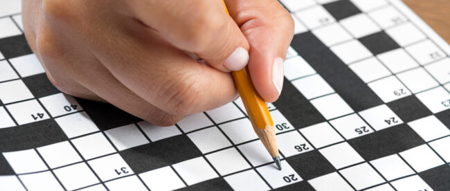 extreme close up of someone's hand. They are holding a pencil and filling up clues on a crossword puzzle.