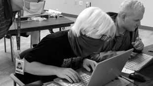 Black and white photo of a student wearing a mask and working on a computer. There is a instructor standing on the side looking at the student’s computer to provide guidance.