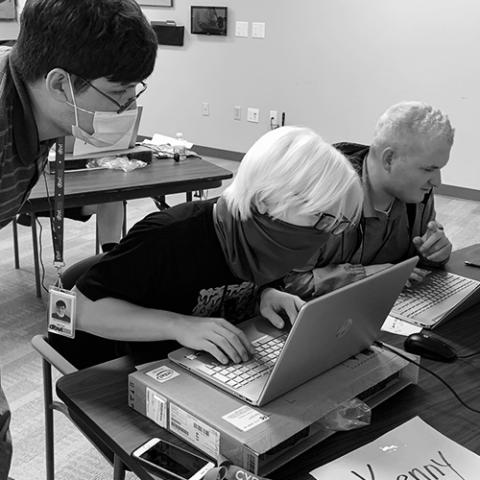 Black and white photo of a student wearing a mask and working on a computer. There is a instructor standing on the side looking at the student’s computer to provide guidance.