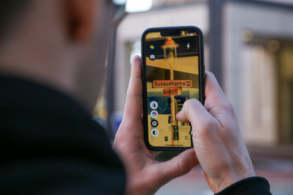 A person using the ReBokeh app to a read street sign. The person is holding a cell phone in and looking at a picture of the street sign on the screen. The street sign is for Susquehanna Avenue W and Washington Ave. 