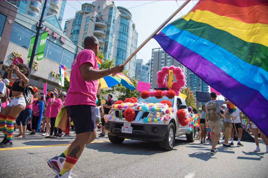 A person wearing a pink shirt and black shorts waving the LGBTQ+ flag at a pride parade.