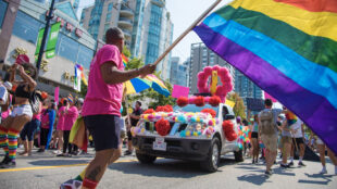 A person wearing a pink shirt and black shorts waving the LGBTQ+ flag at a pride parade.