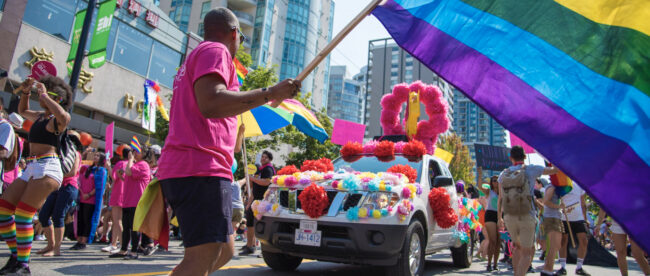 A person wearing a pink shirt and black shorts waving the LGBTQ+ flag at a pride parade.