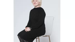 Sugandha Gupta wearing a black dres and black leather shoes, sitting in a white chair and smiling at the camera.