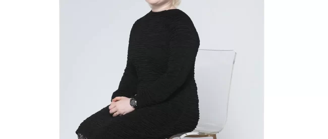 Sugandha Gupta wearing a black dres and black leather shoes, sitting in a white chair and smiling at the camera.