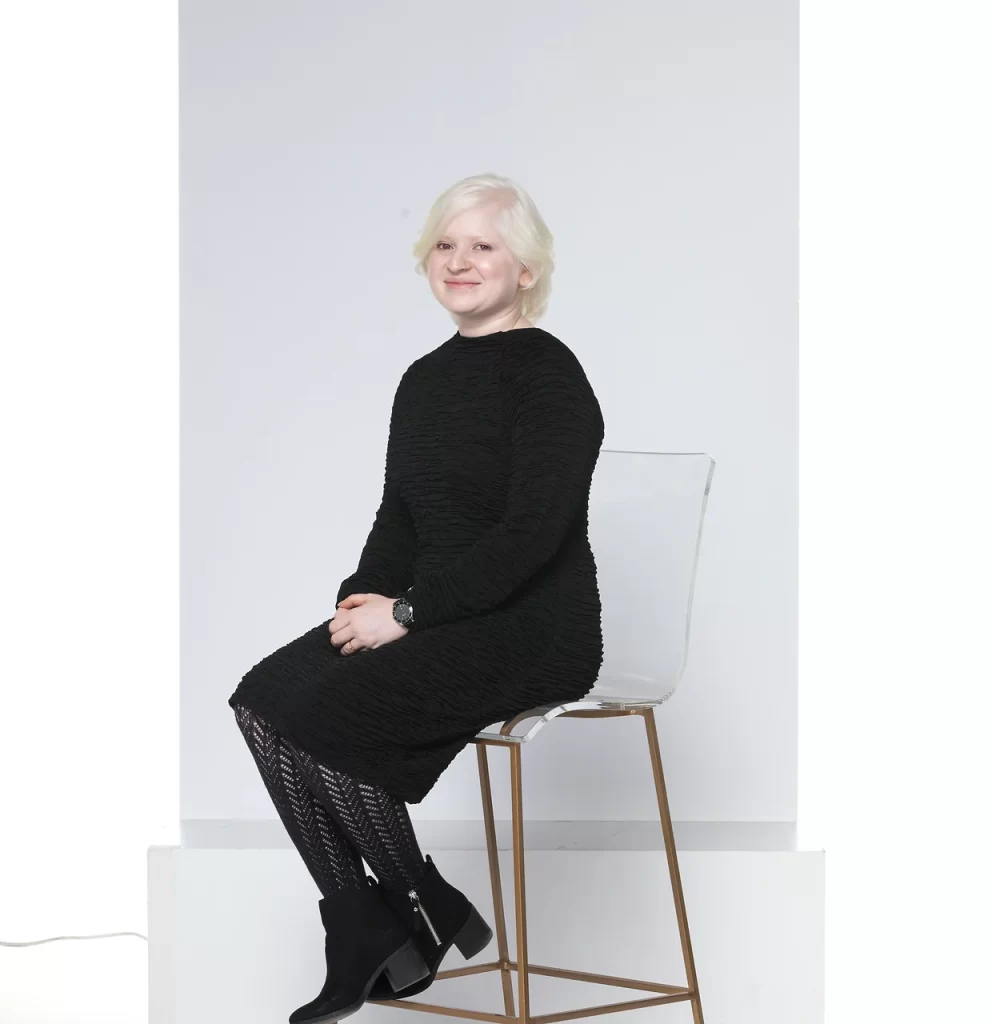 Sugandha Gupta wearing a black dres and black leather shoes, sitting in a white chair and smiling at the camera.