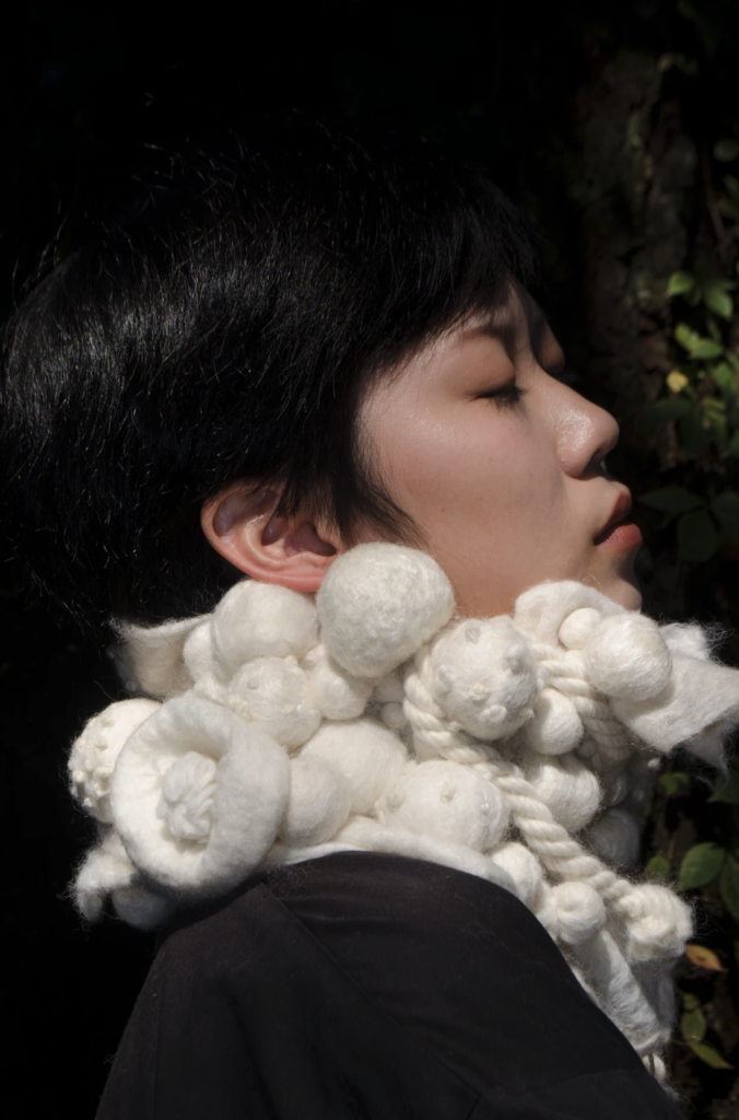 An image with a close up of a sculptural tactile visual wearable piece that can be worn around the neck and shoulder. It is very sculptural and dimensional and has bubble like structure. Each bubble has different materials stuffed inside it such as, beads, cotton balls or fiber. The piece is reversable and is soft and smooth. The bubbles when touching the neck produce many kinds of sensations. The reverse side is not as sculptural but very soft and cushioned. The piece can be dry cleaned for care.