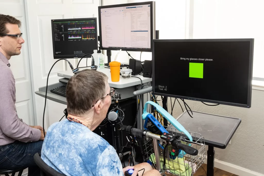 a bespectacled woman with wires coming from an implant in her head looks at a monitor which displays a green square and the word