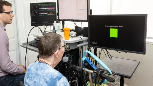 a bespectacled woman with wires coming from an implant in her head looks at a monitor which displays a green square and the word