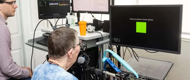 a bespectacled woman with wires coming from an implant in her head looks at a monitor which displays a green square and the word