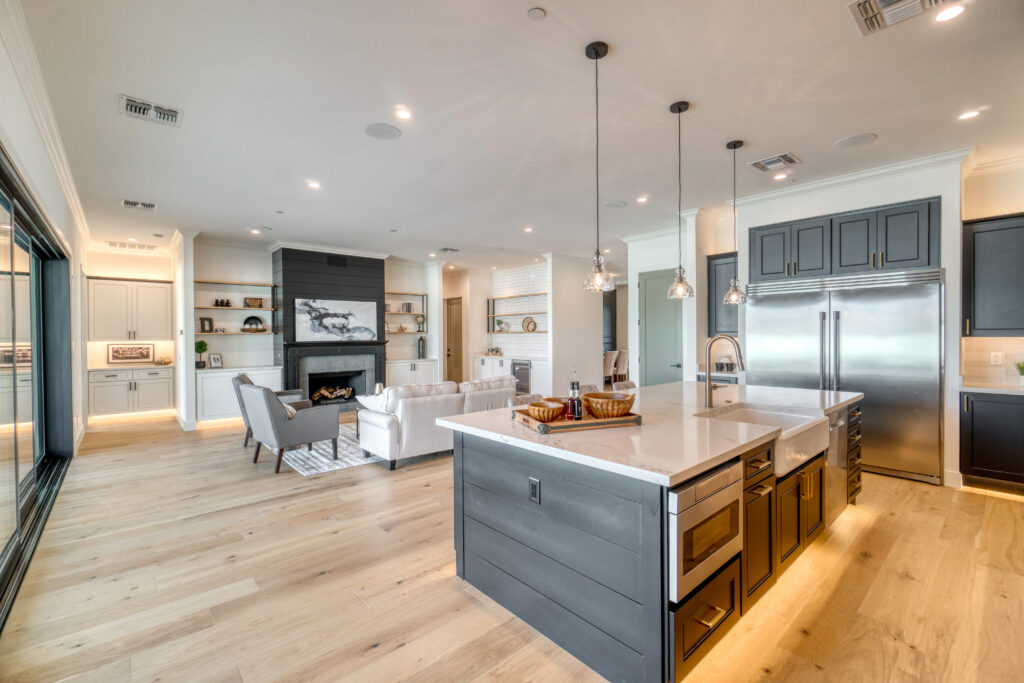 A large, modern kitchen with a large island in the center. The island has a white quartz countertop and stainless steel appliances, including a stovetop, sink, and dishwasher. There are also several bar stools at the island. On the right side of the image is a stainless steel refrigerator and a walk-in pantry. On the left side of the image is a large window overlooking the backyard. The kitchen has white cabinets and light gray walls. There is a hardwood floor throughout the kitchen.