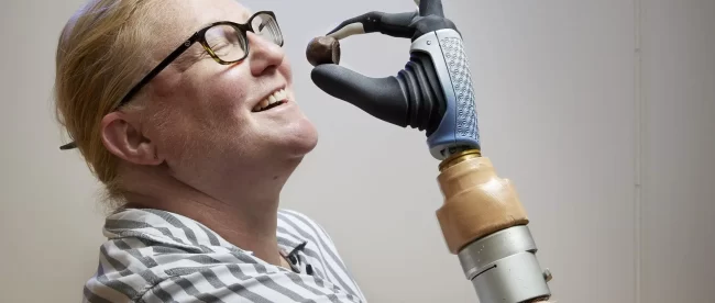 Karin seen holding a small round piece of chocolate with her prosthetic hand close to her mouth and smiling.