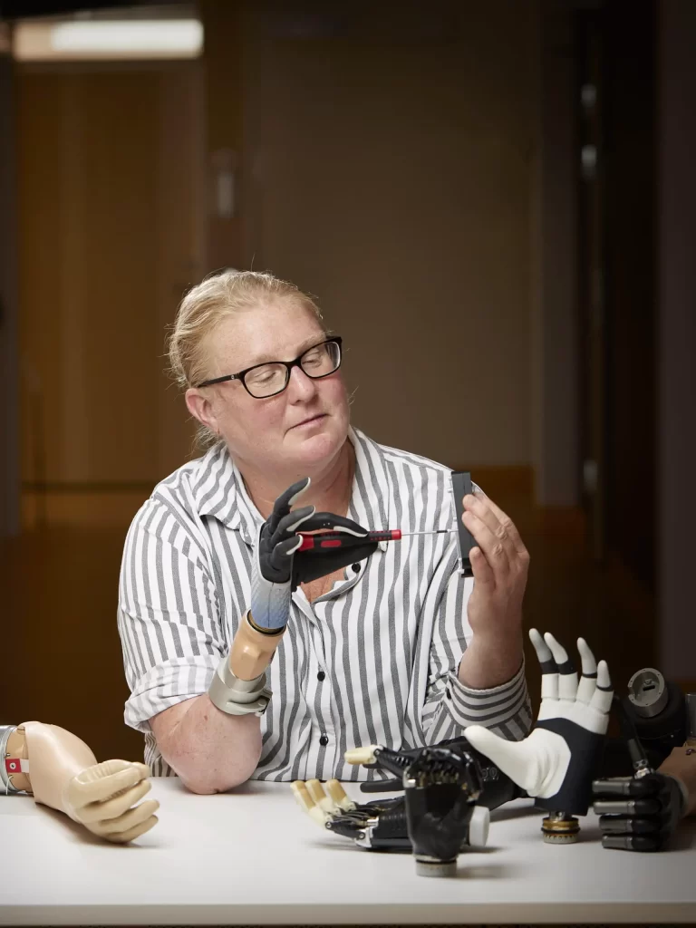 Karin seen using a screwdriver with her prosthetic hand with a gadget. In front of her is another Mia hand resting on the desk.