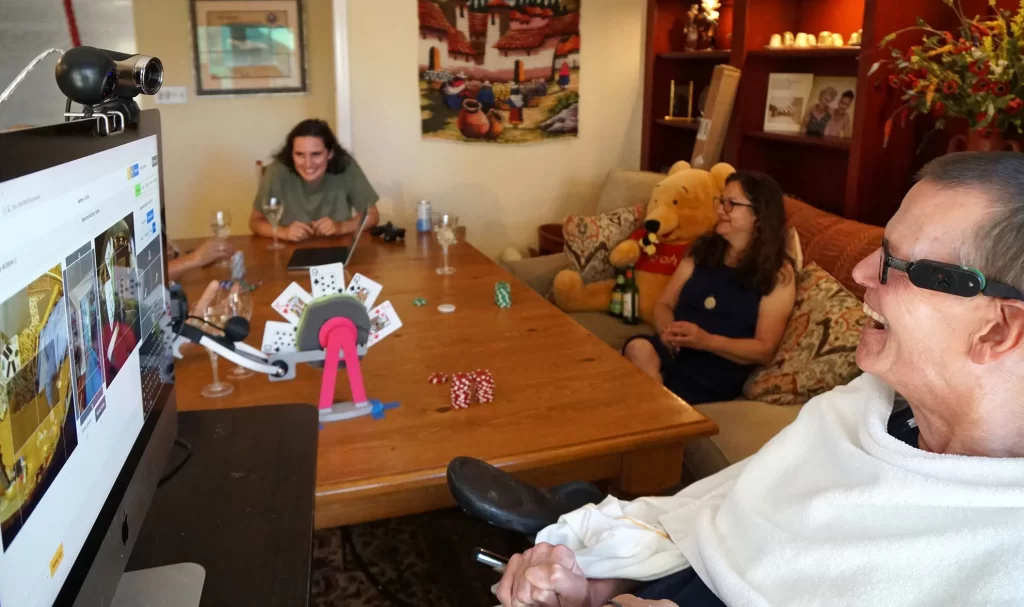 A group of people sit around a table, laughing and playing poker. In the foreground, Henry has a large monitor in front of him showing camera views, as he looks at a device affixed to a robot arm that holds five playing cards.