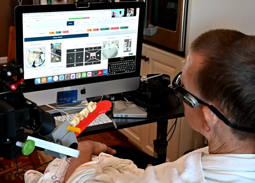 A large monitor shows an interface consisting of multiple views from cameras, simple maps of a house, and a keyboard. Henry is seated in front of the screen, with the arm of a robot just visible, holding a kebab on a red flat tool.