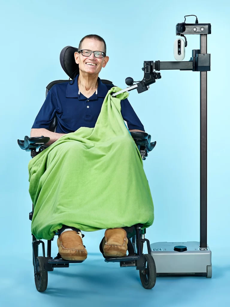 Henry in his wheelchair is seated next to Stretch, the robot, consisting of a mobile base, a thin vertical pole, and a horizontal arm, whose gripper is repositioning a green blanket on him.