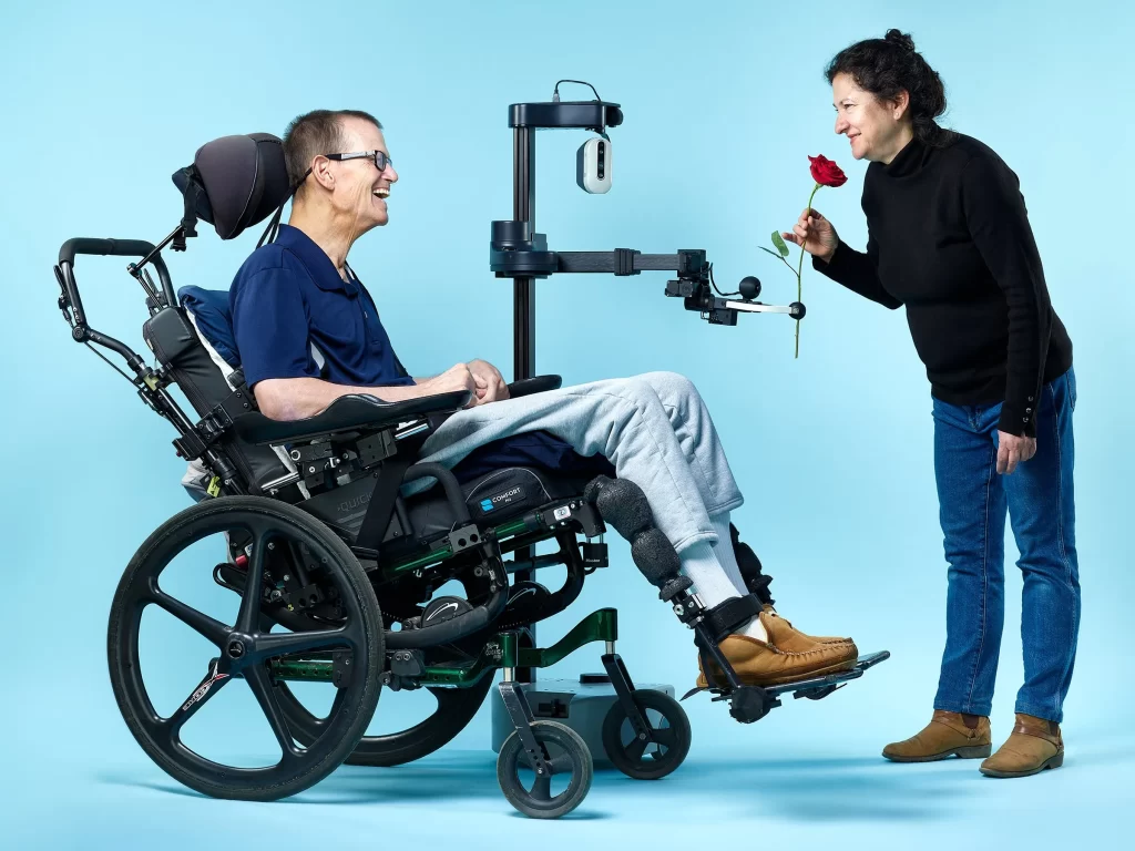 A smiling Henry James in a wheelchair connects eyes with his wife Jane who is in jeans and a turtleneck and bends over to smell a rose that is in the gripper of Stretch.
