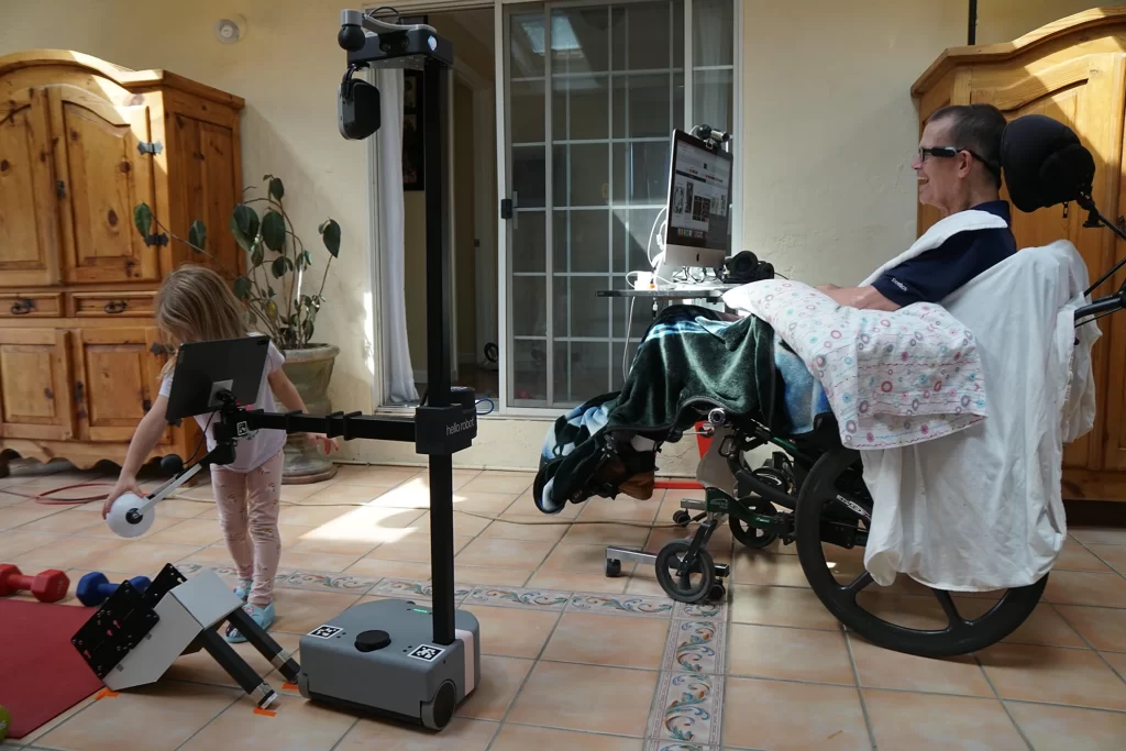 Henry in his wheelchair with a computer is controlling Stretch the robot, which holds a small plastic bowling ball that a small child is also grabbing onto.