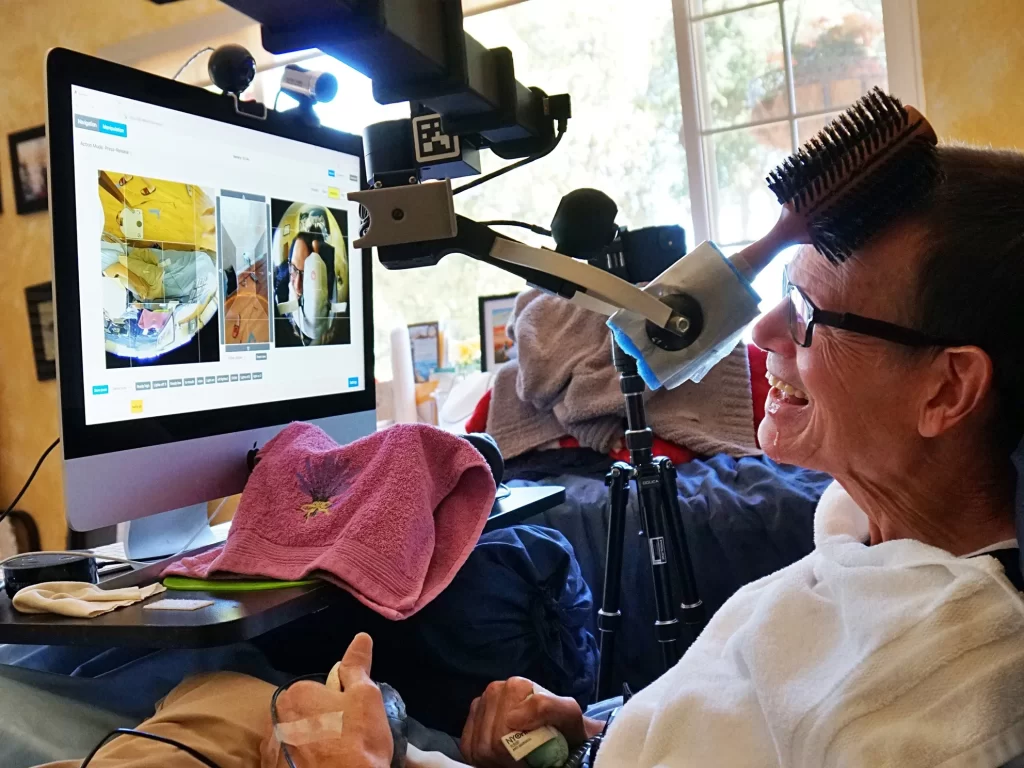 Henry lies in bed. He is looking at a monitor which shows multiple camera views, including one of himself, as a robotic gripper holding a hairbrush scratches his head.