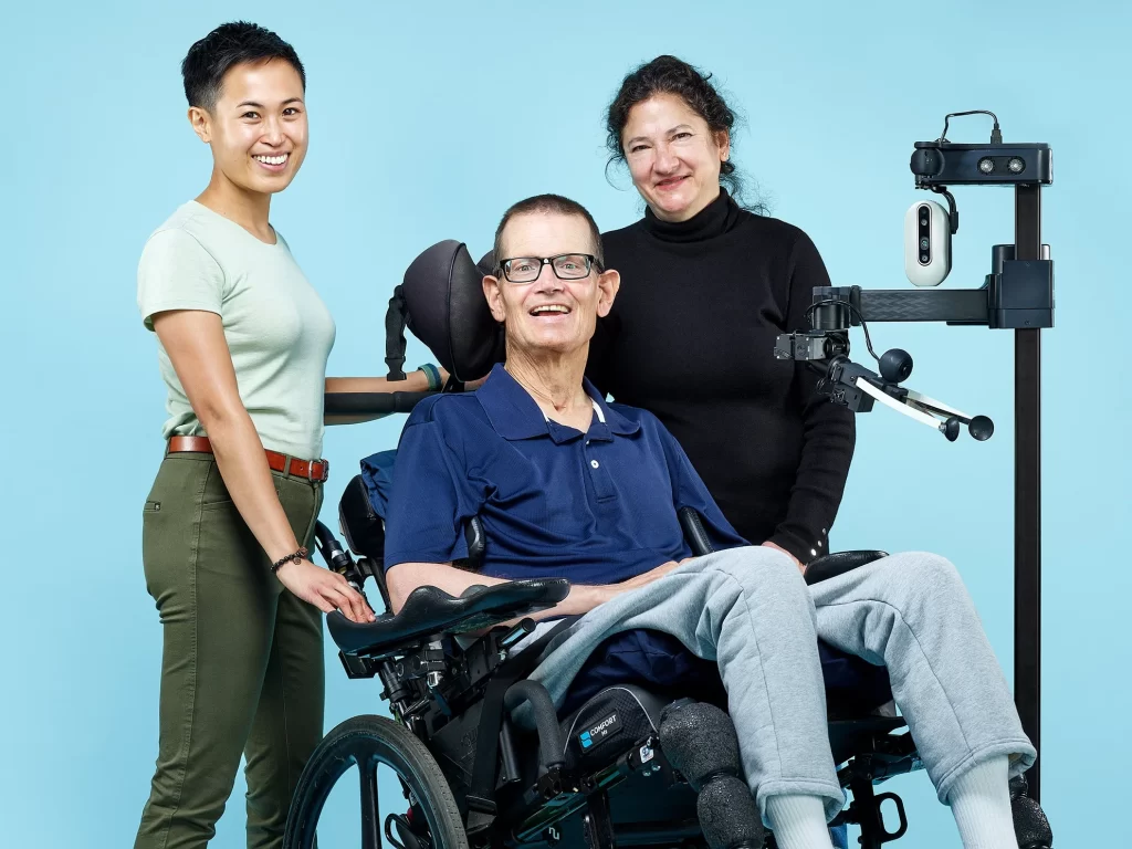 Group portrait of Vy Nguyen, an occupational therapist who has short hair and is smiling, henry in his wheelchair smiling, a smiling Jane in a black turtleneck, and Stretch the robot.