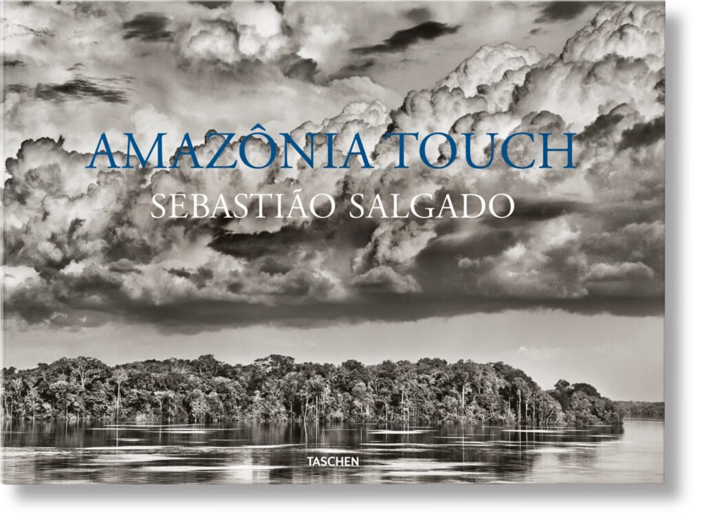 A black and white photo of a small island in the middle of a large body of water surrounded by clouds. The text Amazon Touch Sebastian Salgado is laid on top of the image.