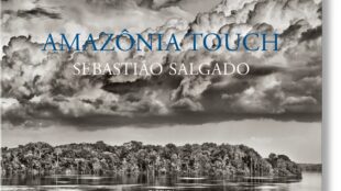 A black and white photo of a small island in the middle of a large body of water surrounded by clouds. The text Amazon Touch Sebastian Salgado is laid on top of the image.