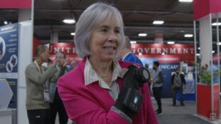 The image features an older woman with short grey hair, smiling. She is wearing a bright pink jacket and is GyroGlove. In the background, there are people and booths at CES2024.