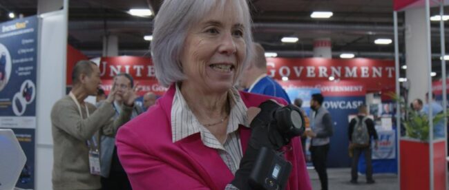 The image features an older woman with short grey hair, smiling. She is wearing a bright pink jacket and is GyroGlove. In the background, there are people and booths at CES2024.