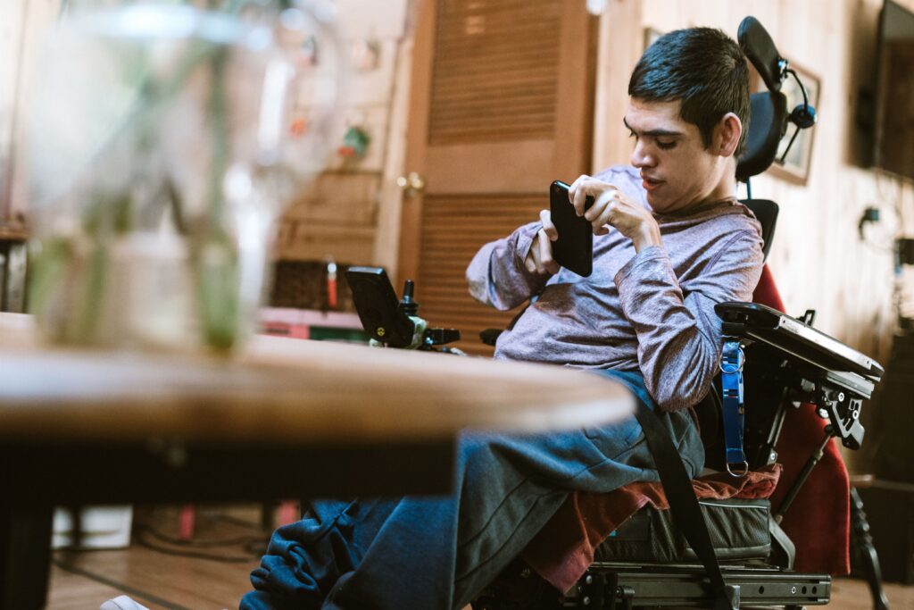 an adult man in a power wheelchair engaging with a smartphone. He appears focused on the device, which he is holding with his right hand. The setting looks like an indoor environment, possibly a home, with a blurred background that suggests a casual, lived-in space. The man's left hand rests on the wheelchair's control interface, and his posture and expression indicate concentration or communication.