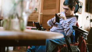 an adult man in a power wheelchair engaging with a smartphone. He appears focused on the device, which he is holding with his right hand. The setting looks like an indoor environment, possibly a home, with a blurred background that suggests a casual, lived-in space. The man's left hand rests on the wheelchair's control interface, and his posture and expression indicate concentration or communication.