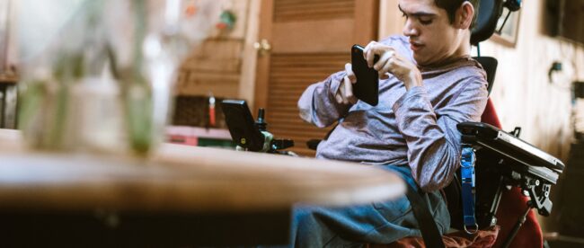 an adult man in a power wheelchair engaging with a smartphone. He appears focused on the device, which he is holding with his right hand. The setting looks like an indoor environment, possibly a home, with a blurred background that suggests a casual, lived-in space. The man's left hand rests on the wheelchair's control interface, and his posture and expression indicate concentration or communication.