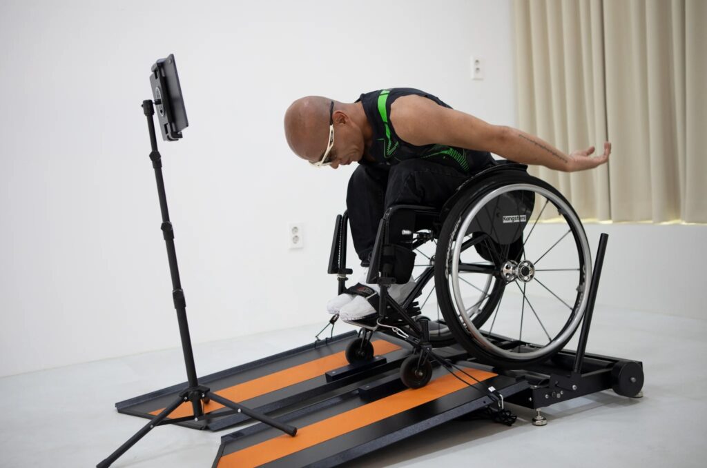 The image features an individual in a wheelchair using a specialized treadmill designed for wheelchair users, called the Wheely-X. The user, who is bald and wearing glasses along with a sleeveless sports top, is in motion, with one arm extended backward, suggesting dynamic movement. In front of him, there is a tablet mounted on a stand, likely displaying an app or game that interacts with the treadmill. The treadmill itself has bright orange stripes on a dark surface, which contrasts with the minimalist and bright background of the room. This setup appears to be designed for both exercise and entertainment, potentially for use in a home or a rehabilitation center.