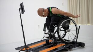 The image features an individual in a wheelchair using a specialized treadmill designed for wheelchair users, called the Wheely-X. The user, who is bald and wearing glasses along with a sleeveless sports top, is in motion, with one arm extended backward, suggesting dynamic movement. In front of him, there is a tablet mounted on a stand, likely displaying an app or game that interacts with the treadmill. The treadmill itself has bright orange stripes on a dark surface, which contrasts with the minimalist and bright background of the room. This setup appears to be designed for both exercise and entertainment, potentially for use in a home or a rehabilitation center.