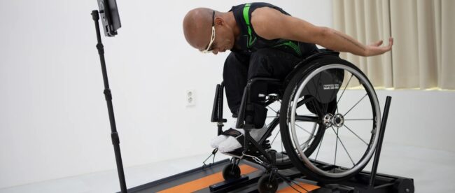 The image features an individual in a wheelchair using a specialized treadmill designed for wheelchair users, called the Wheely-X. The user, who is bald and wearing glasses along with a sleeveless sports top, is in motion, with one arm extended backward, suggesting dynamic movement. In front of him, there is a tablet mounted on a stand, likely displaying an app or game that interacts with the treadmill. The treadmill itself has bright orange stripes on a dark surface, which contrasts with the minimalist and bright background of the room. This setup appears to be designed for both exercise and entertainment, potentially for use in a home or a rehabilitation center.