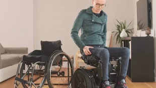 A person is sitting on the Kim-e, which is positioned next to the traditional wheelchair for comparison. Kim-e features a sleek design and a pair of large wheels, similar to those found on a Segway, which suggests its self-balancing capabilities. The person is wearing casual attire and is looking down. The environment suggests an indoor setting, possibly a living room, indicating the practical use of the Kim-e in everyday life at home.