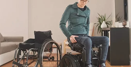 A person is sitting on the Kim-e, which is positioned next to the traditional wheelchair for comparison. Kim-e features a sleek design and a pair of large wheels, similar to those found on a Segway, which suggests its self-balancing capabilities. The person is wearing casual attire and is looking down. The environment suggests an indoor setting, possibly a living room, indicating the practical use of the Kim-e in everyday life at home.