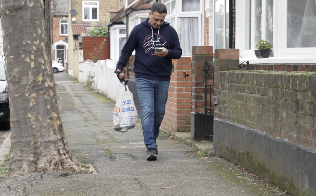 The image shows Suleman Chohan walking down a residential sidewalk, looking at his smartphone. He is dressed casually in a hoodie and jeans and is carrying a plastic shopping bag in his right hand. The surroundings suggest an overcast day in a suburban neighborhood with houses and parked cars in the background. He is using the hero gauntlet on his right arm to grip grocery bags.